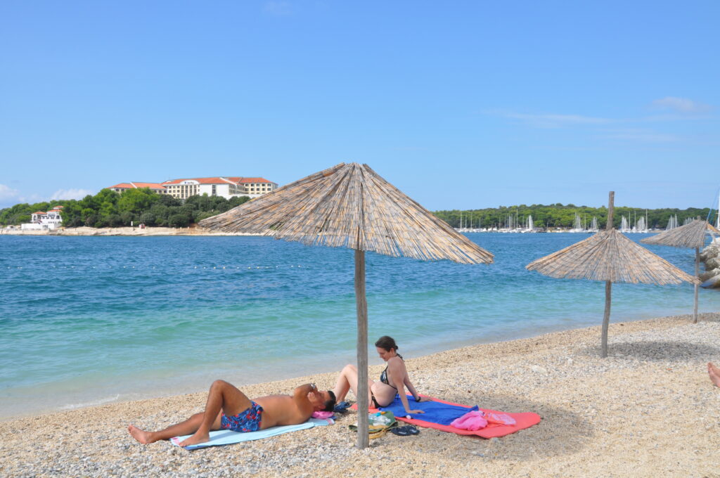 Żwirowa plaża na Istrii z turkusowym morzem i trzcinowymi parasolami